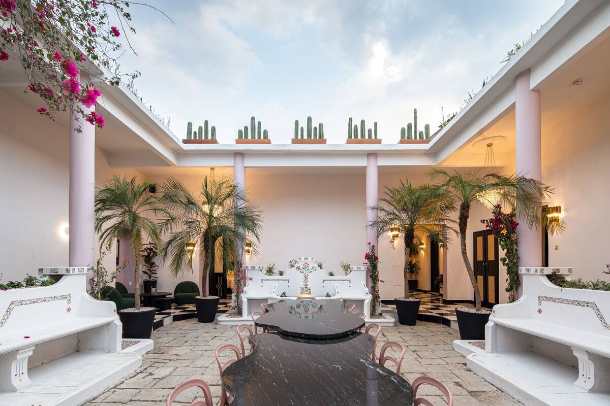 Un vaste patio intérieur doté d’une table ronde centrale, entouré de bancs blancs et de plantes en pot.
