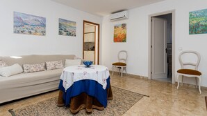 Ein Wohnzimmer mit einer Couch, einem Esstisch mit blauer Tischdecke, zwei Stühlen und Bildern an den Wänden.