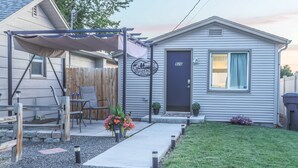 Une petite maison de campagne à un seul étage, avec une porte d’entrée sombre, un petit porche doté d’un espace salon abrité et une pelouse bien entretenue.