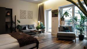 Een moderne woonkamer met een bed, bank en fauteuil, houten vloeren en grote ramen met planten.