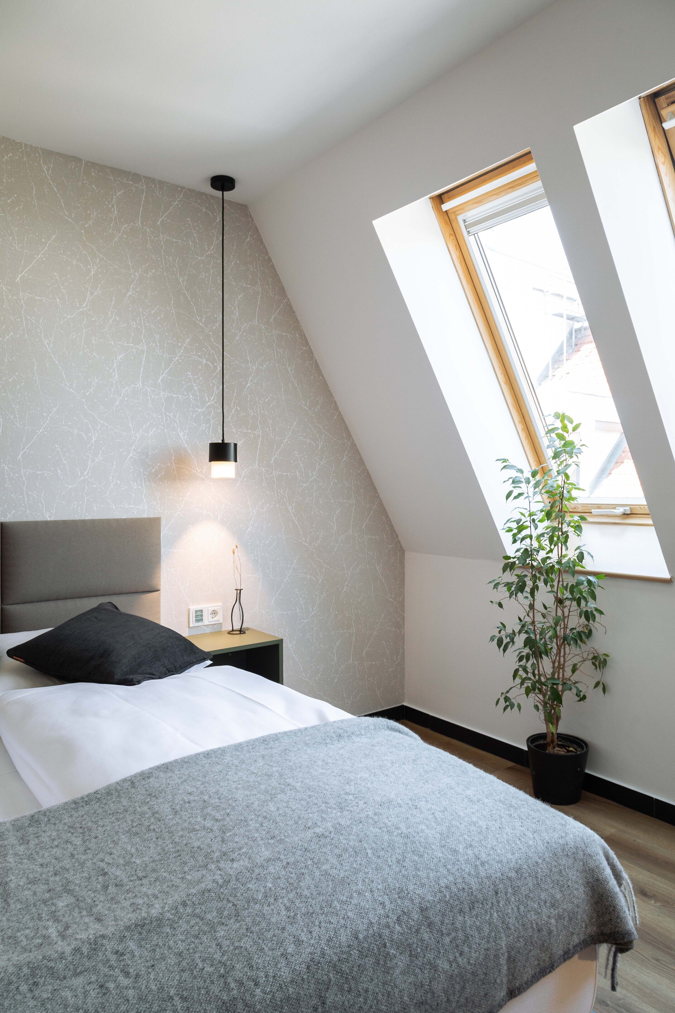 A modern bedroom with a sloped ceiling, a skylight, a bedside table with a lamp, a bed with a grey blanket, and a potted plant.