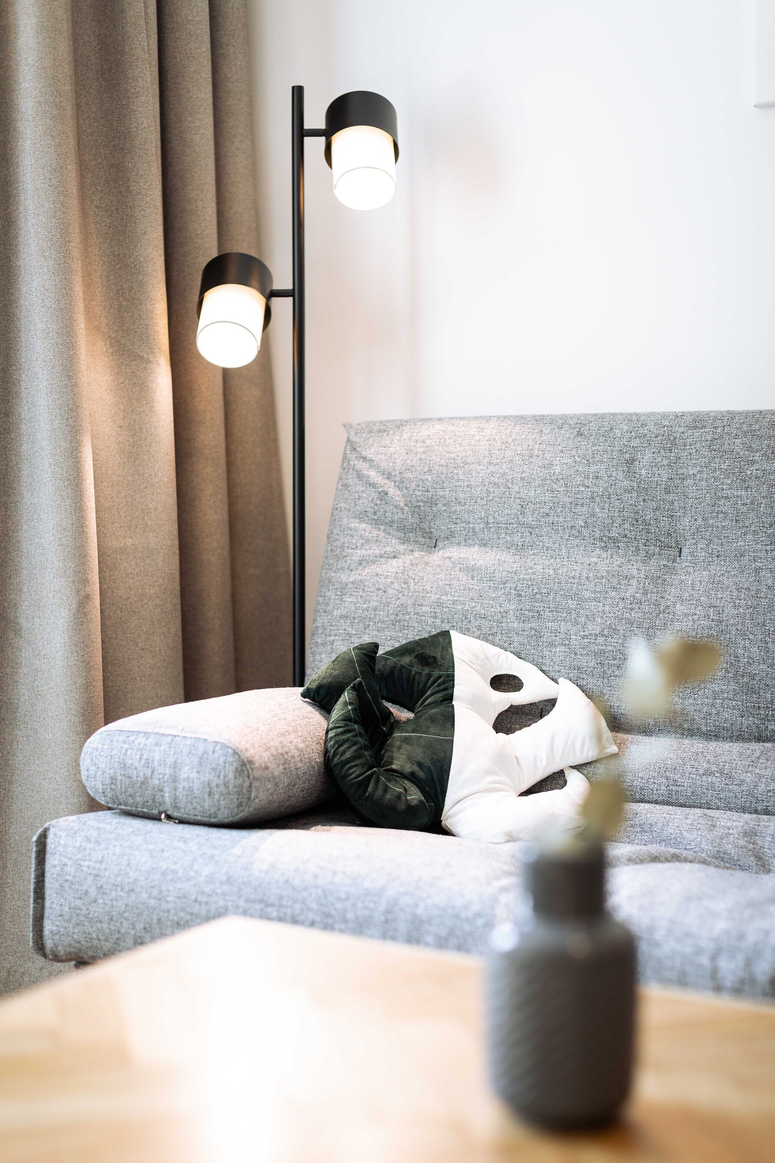 Een moderne woonkamer met een grijze bank, een zwarte vloerlamp en een vaas op een houten tafel.