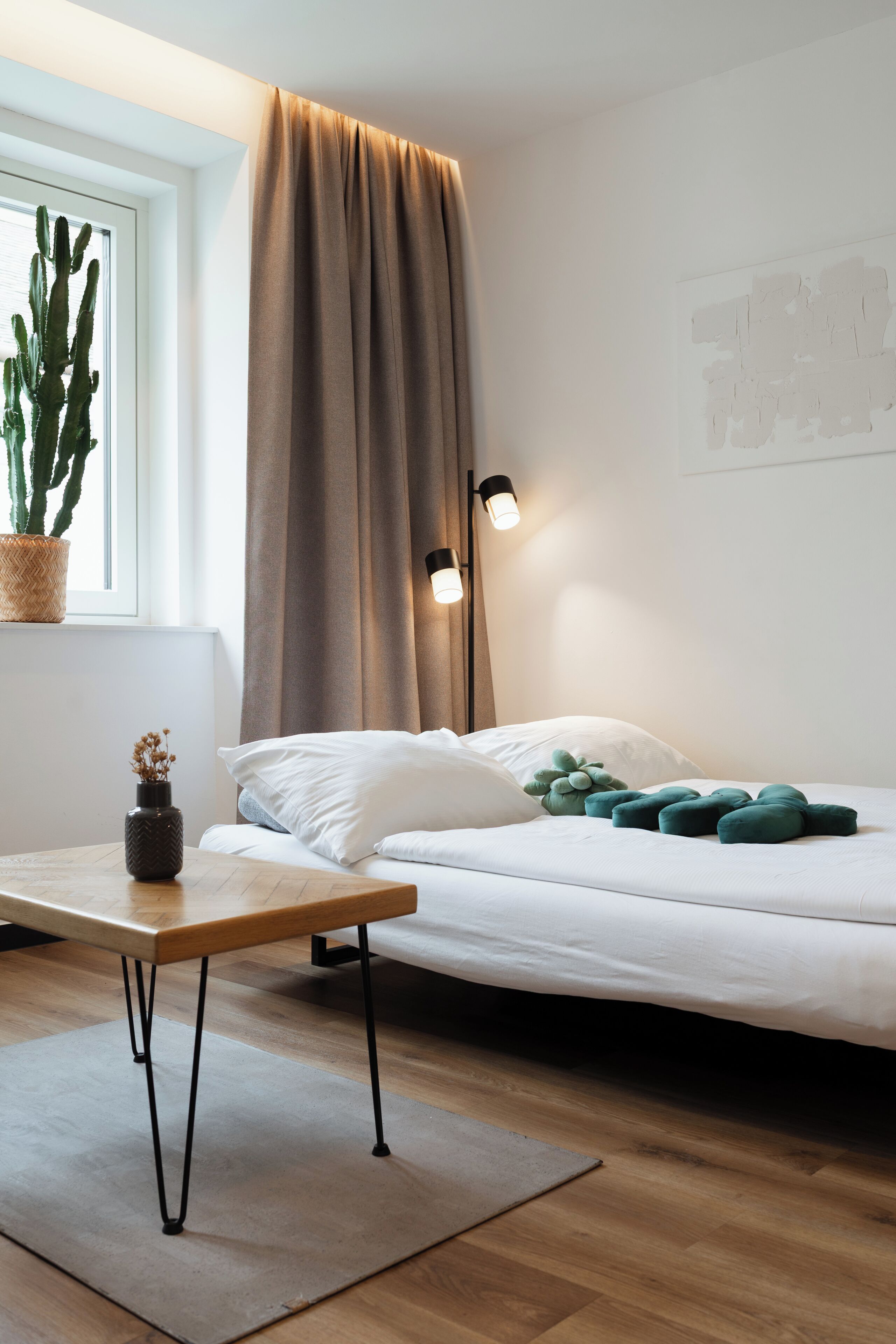 A bedroom with a bed, a nightstand, a window with a cactus, and a wall-mounted lamp.