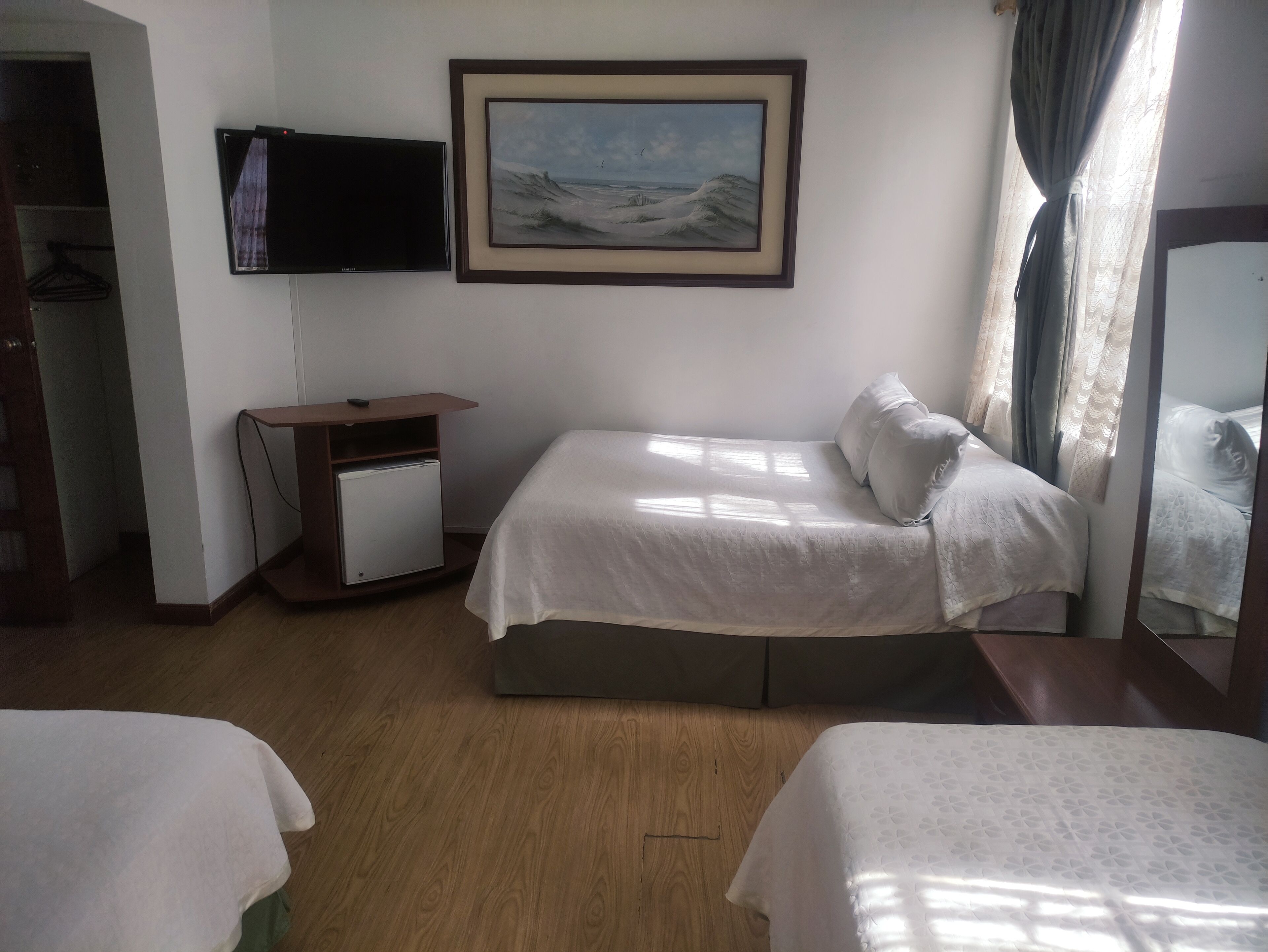 A hotel room with a bed, a small desk with a refrigerator, a TV mounted on the wall, and a painting above the bed.