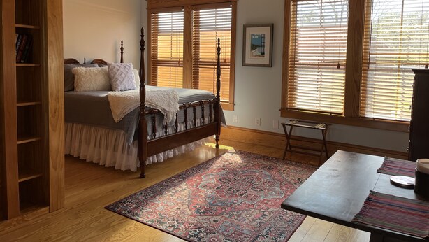 Un dormitorio con una cama de madera, una mesita de noche, una estantería y ventanas amplias con persianas.