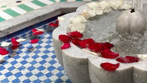 Uma fonte de mármore com água jorrando do centro, rodeada por pétalas de rosa vermelhas.