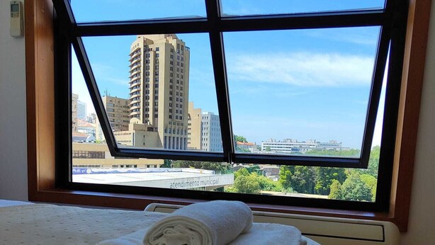 Une chambre d’hôtel dotée d’une grande fenêtre donnant sur la ville, d’un lit bien fait avec du linge de maison blanc et d’un appareil de climatisation visible.