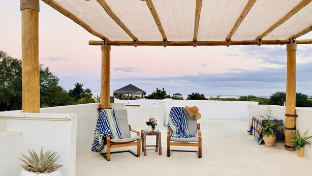 Un espace extérieur abrité, aménagé avec deux chaises en bois et une petite table, d’où l’on peut admirer une plage avec, en arrière-plan, des constructions aux toits de chaume.