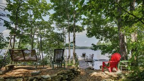 A lakeside deck with a swing chair, a rocking chair, and a red Adirondack chair. - Maine Cottage w/ Dock & Kayaks, Near Augusta! (Augusta)
