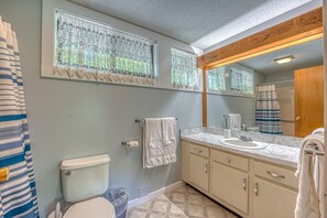 Ein Badezimmer mit Toilette, Waschbecken und Waschtisch. An den Fenstern hängen Spitzenvorhänge, und ein Handtuch ist an der Wand aufgehängt.