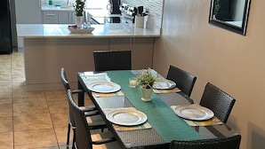 Un coin repas avec une longue table dressée pour six personnes, entourée de chaises en osier. On aperçoit une cuisine à l’arrière-plan, avec une fenêtre donnant sur un espace verdoyant.