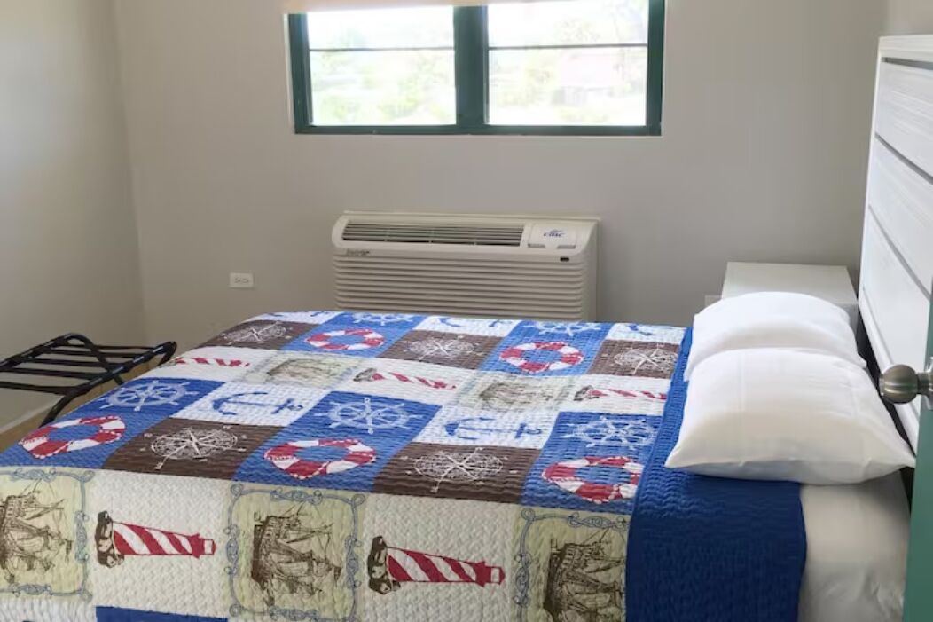 Un dormitorio con colcha acolchada, una ventana con vistas y aire acondicionado de pared, además de un aparador blanco.