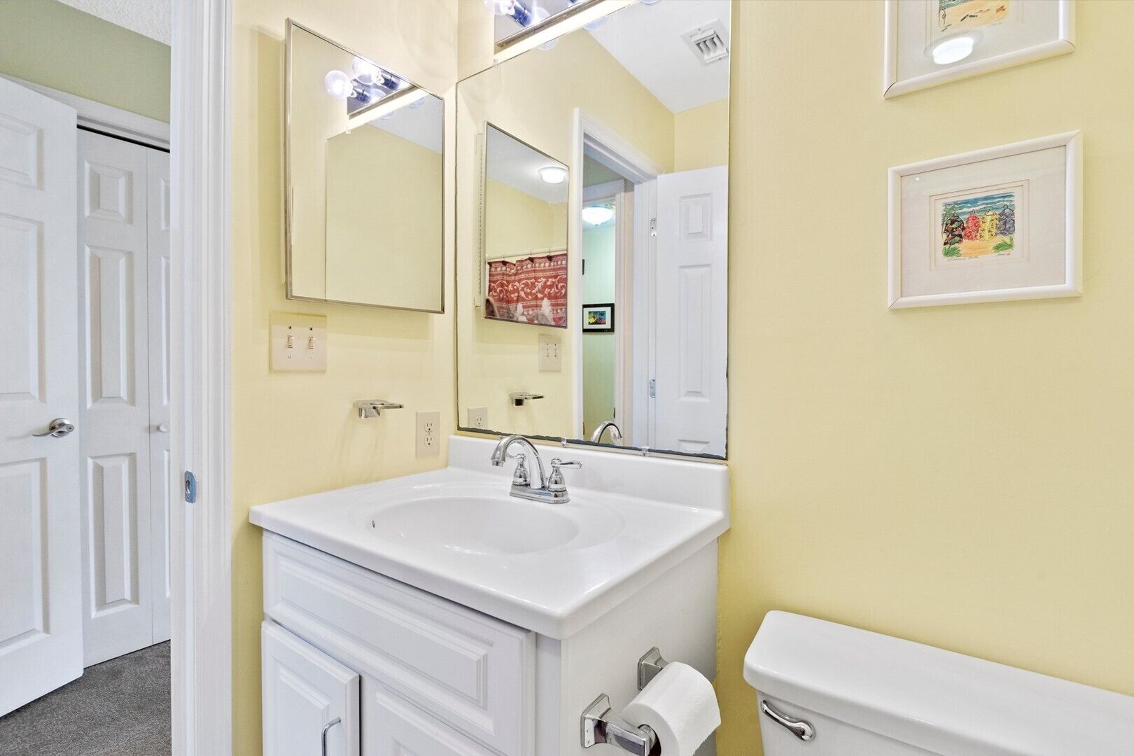 A bathroom with a white sink, mirror, and toilet. There are framed pictures on the wall and a door to the left.
