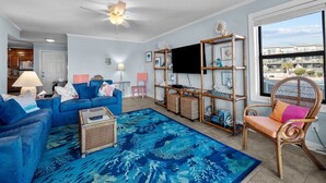 Ein Wohnzimmer mit blauem Sofa, einem Couchtisch, einem blau gemusterten Teppich, einem Fernseher und einem Fenster mit Blick auf Gebäude.