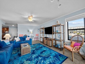 Un salon avec un canapé bleu, une table basse, un tapis bleu à motifs, un téléviseur et une fenêtre donnant sur des immeubles.