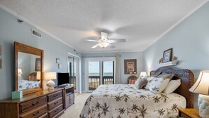 Ein Schlafzimmer mit Bett, Kommode, Deckenventilator und Blick auf den Ozean durch die Balkontüren.