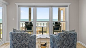 A room with a view of the beach, two Adirondack chairs, a small table, and a wooden floor. - 3BR Oceanfront Condo with Community Pool (North Topsail Beach)