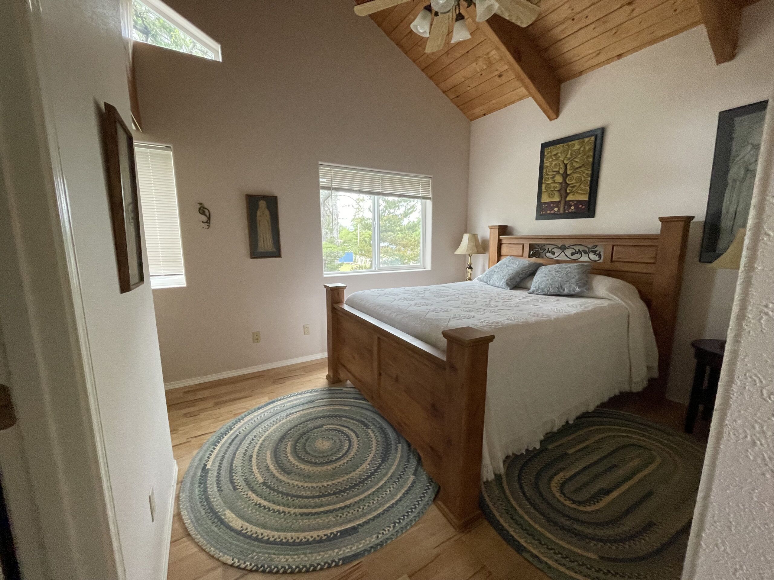 Une chambre à coucher avec un lit en bois, deux fenêtres, un ventilateur de plafond et un tableau accroché au mur.