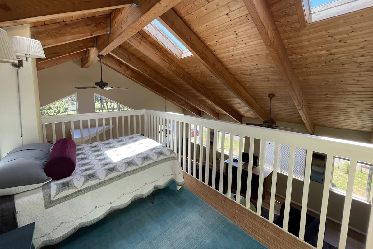 A bedroom with a wooden ceiling, a bed with a floral bedspread, a white railing, and a view of the outdoors.