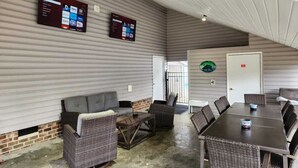 A covered outdoor area with a long table and chairs, a sofa, and two mounted screens displaying content.