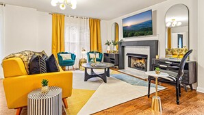 A living room with a yellow sofa, teal armchairs, a fireplace, and a large TV. - Pet-Friendly, Sauna, Hot Tub, Game Room, Two Firepits, Sleeps 12 (Asheville)