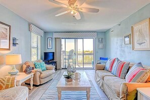 A living room with a ceiling fan, a TV, a sofa, a coffee table, and a view through the glass door. - 2BR Oceanfront Condo with Community Pool & Elevator (North Topsail Beach)