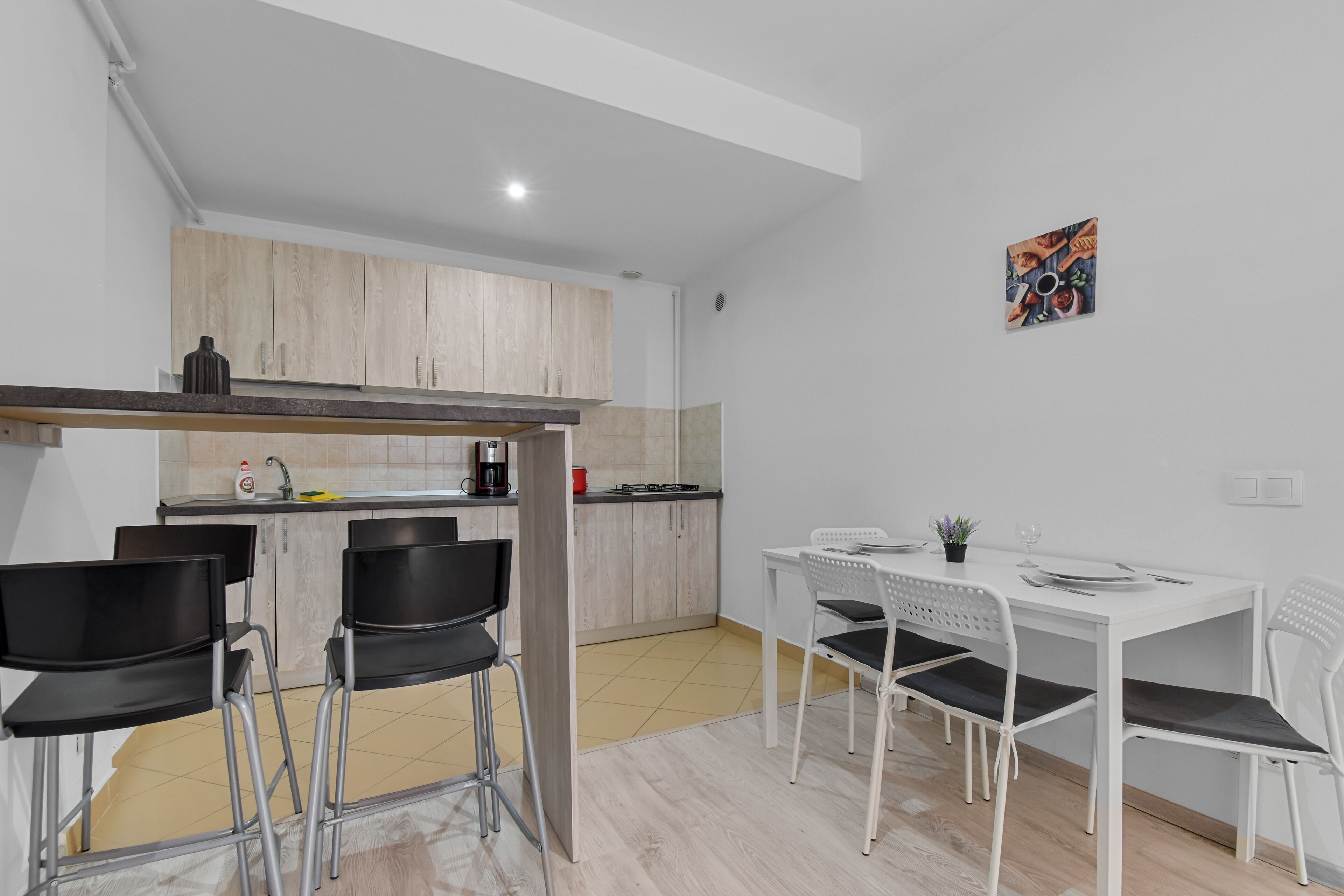 Cocina compacta con comedor, que incluye una mesa y sillas blancas, una cafetera y un mueble pequeño.