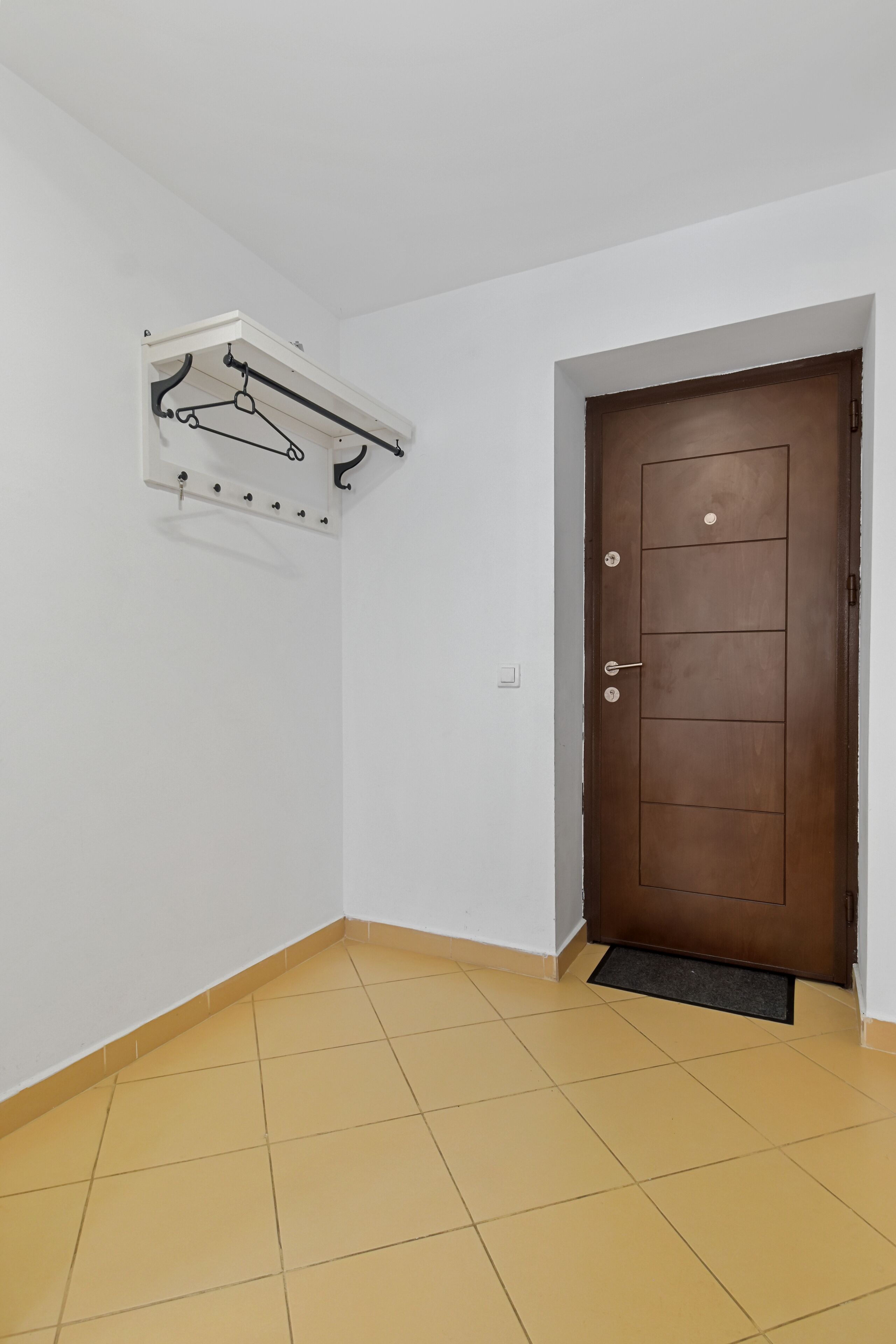 Una habitación con una puerta marrón, un perchero blanco fijado a la pared y un suelo de baldosas amarillas.