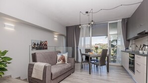 Un appartement moderne avec un canapé gris, une table à manger et des chaises. Il comprend un coin cuisine avec des placards et offre une vue sur la ville grâce à de grandes fenêtres.