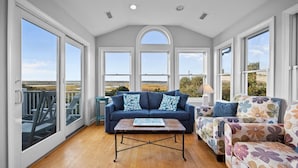 A spacious room with a high ceiling, large windows, a blue sofa, a floral armchair, and a wooden coffee table. - 5BR Ocean View House on North Topsail Beach - Pool (North Topsail Beach)