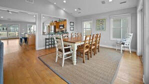 A spacious dining area with a wooden table and chairs, a kitchen in the background, and a view of the outside deck. - 5BR Ocean View House on North Topsail Beach - Pool (North Topsail Beach)