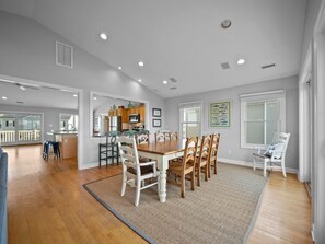 A spacious dining area with a wooden table and chairs, a kitchen in the background, and a view of the outside deck. - 5BR Ocean View House on North Topsail Beach - Pool (North Topsail Beach)