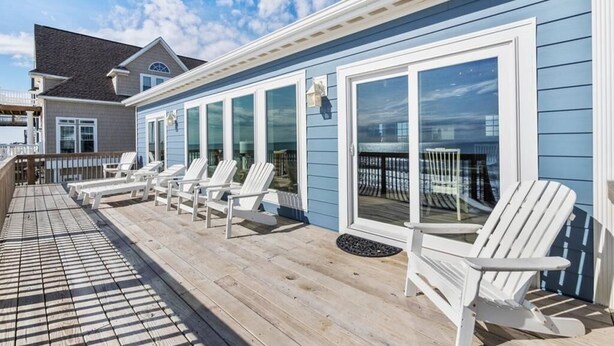 A wooden deck with white Adirondack chairs, a glass-enclosed patio area, and a view of the ocean.
