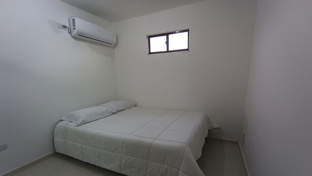 Una habitación pequeña con cama individual, paredes blancas, ventana y un equipo de aire acondicionado en la pared.