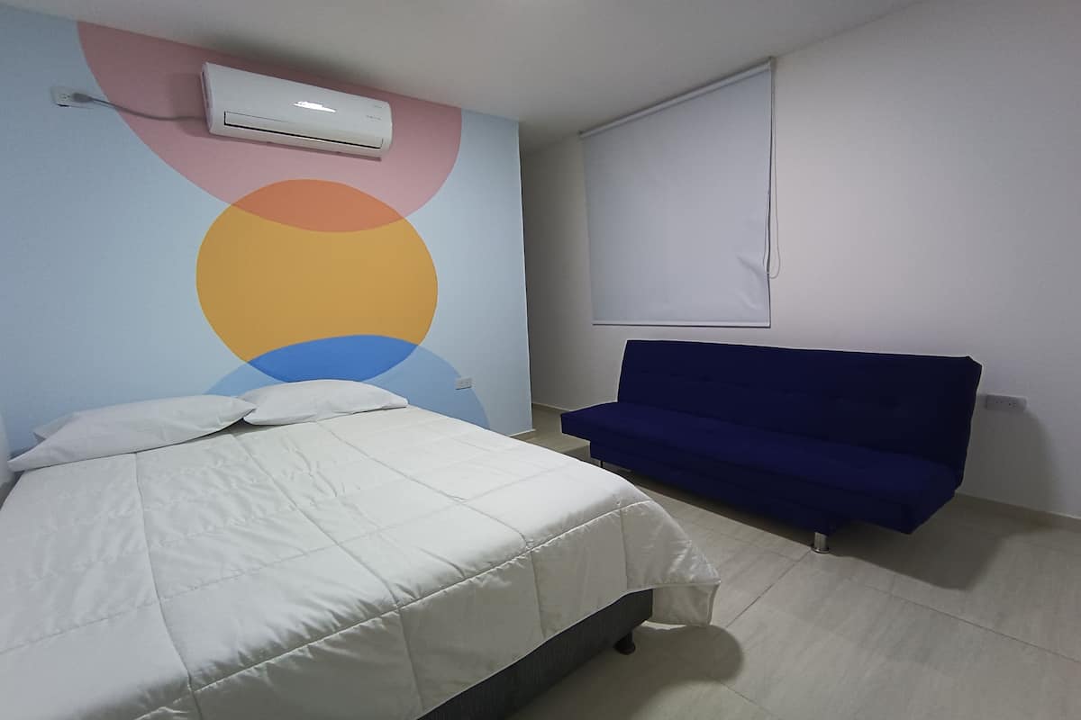 A bedroom with a bed, a blue sofa, a wall with a colorful circular design, and an air conditioning unit.