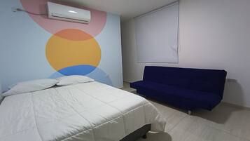 Une chambre à coucher avec un lit, un canapé bleu, un mur orné d’un motif circulaire coloré et un appareil de climatisation.