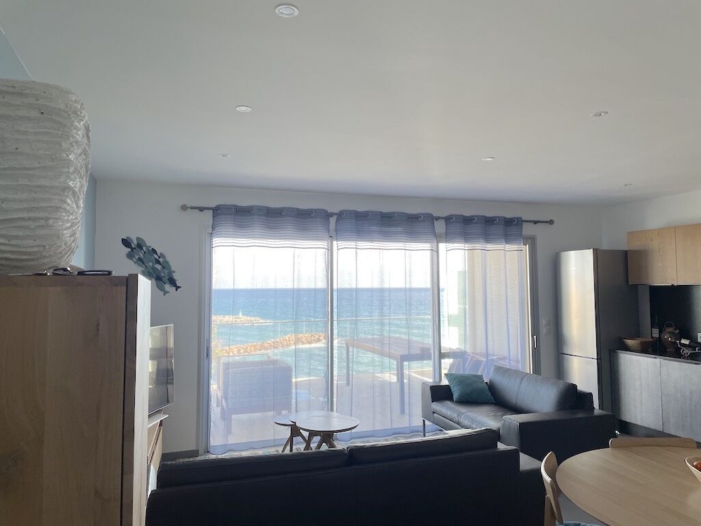 A modern living room with a large sliding glass door leading to a balcony with a view of the sea. There is a sofa, a small round table, and a dining area with a wooden table and chairs.