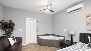 A modern bedroom with a large bed, a sitting area with a couch, a small side table, a wall-mounted air conditioner, a ceiling fan, and a large window. - Gorgeous New 1 bd condo - 5 Min to Branson Landing (Branson)