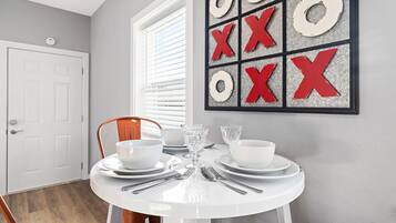 A dining area with a round table set for two, white plates, bowls, and cutlery. A red and white decorative piece hangs on the wall.