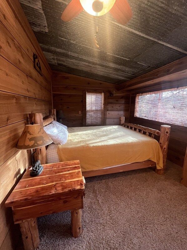 Een houten kamer met een bed, een lamp, een klein tafeltje en een plafondventilator.
