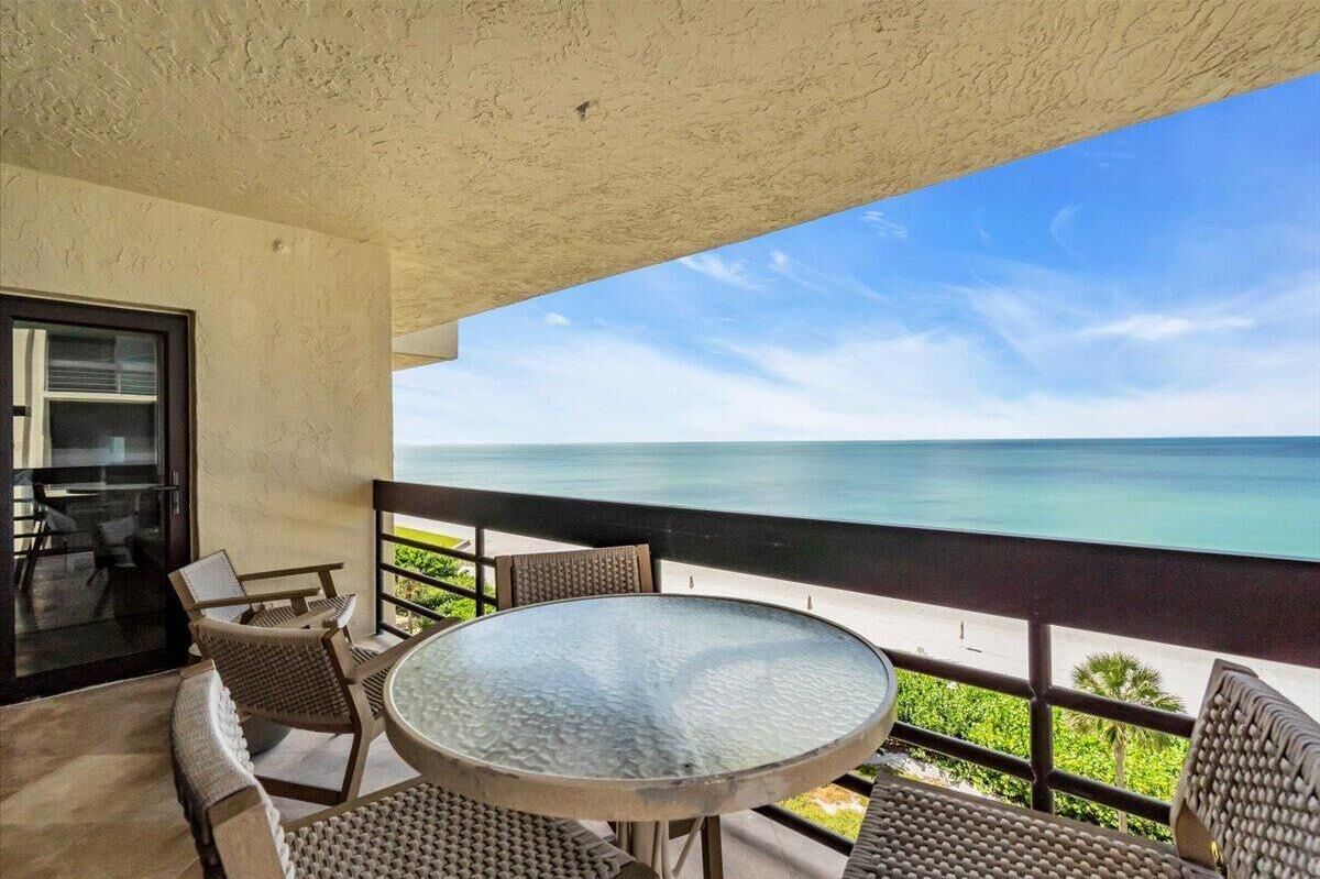 Een balkon met een glazen tafel en rieten stoelen, met uitzicht op een strand.