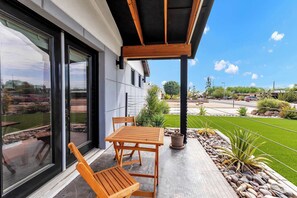 A modern outdoor patio with a wooden table and chairs, a potted plant, and a view of a landscaped garden and street.