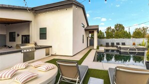 Un espace cuisine extérieur moderne, doté d’un barbecue, d’une piscine et de coins salon avec des coussins.