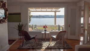 Een kamer met uitzicht op een waterpartij, twee stoelen, een kleine tafel met een vaas en een bloempot.