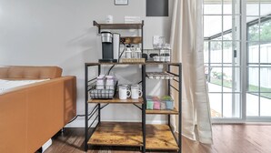 Een kamer met een bank, een koffiehoek met koffiezetapparaat, kopjes en een bord met koffiesoorten.
