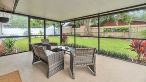Een overdekt terras met rieten meubels en een glazen tafel, met uitzicht op een achtertuin met een houten schutting en groen.