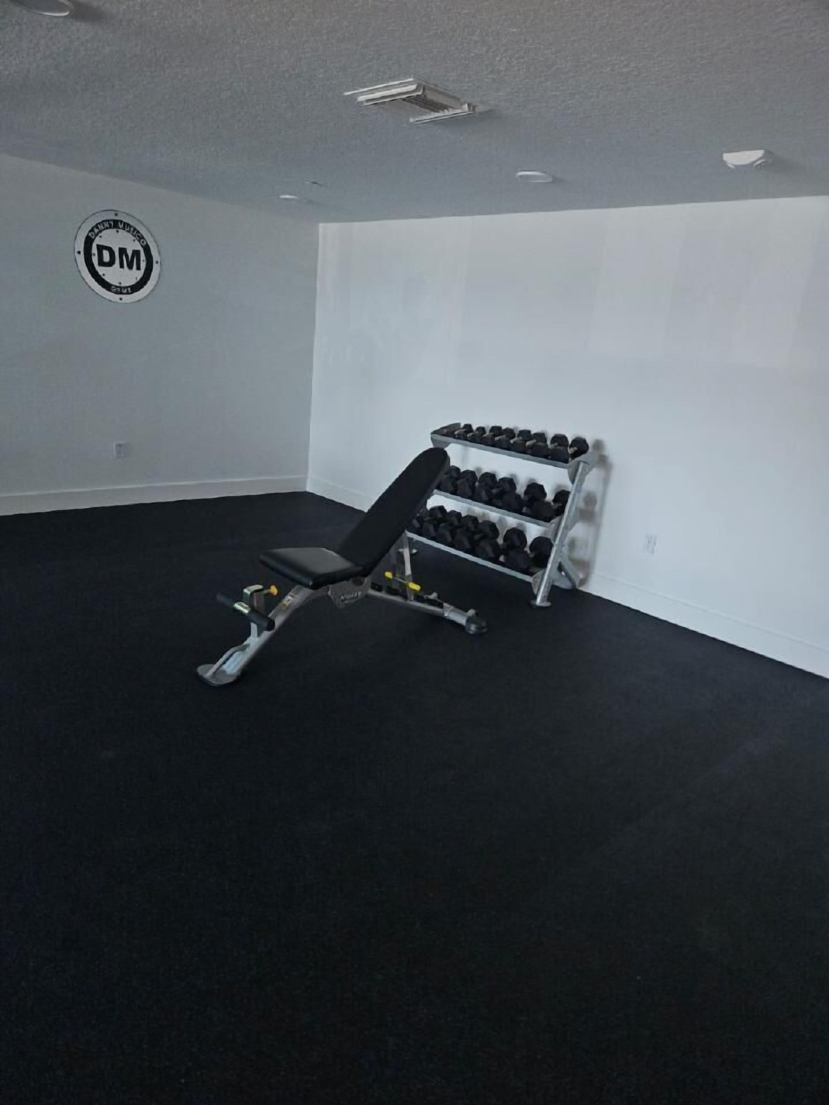 Ein Fitnessraum mit einer Langhantelbank, einem Hantelständer und einem Logo an der Wand.