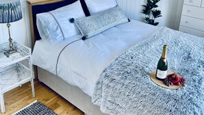 Cama bem arrumada com travesseiro azul e branco estampado, cabeceira de madeira e mesa de cabeceira com abajur. Uma garrafa de champanhe e morangos estão sobre uma bandeja de madeira redonda na cama.