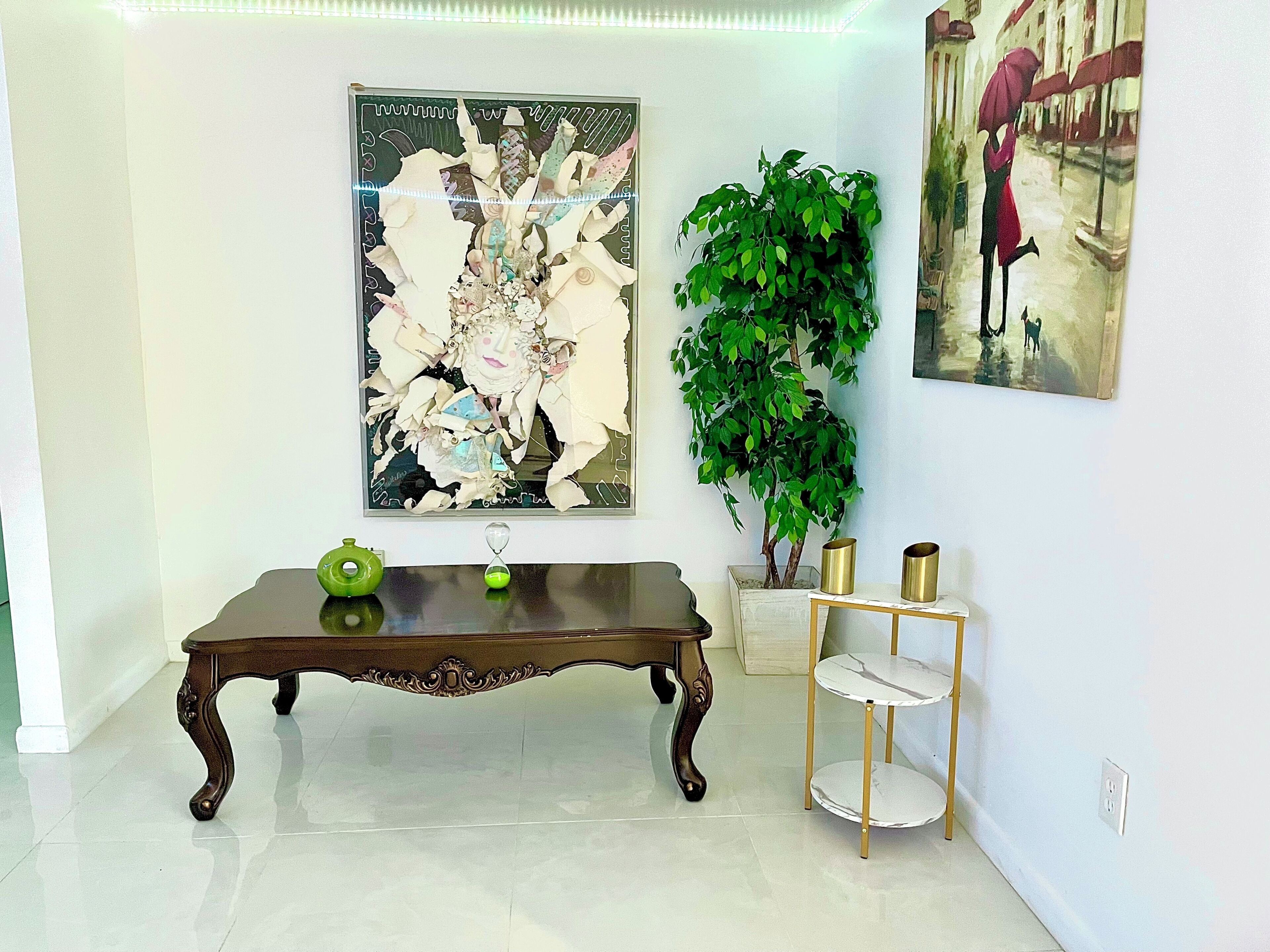 A room with a wooden table, a green decorative item, a wall-mounted painting, a potted plant, and a gold-framed shelf.