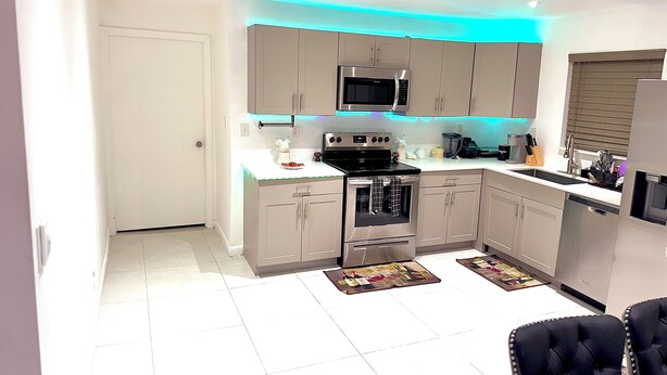 Een moderne keuken met grijze kastjes, een zwarte magnetron, een fornuis en een spoelbak. Er liggen twee vloerkleden op de vloer en er staat een zwarte stoel op de voorgrond.